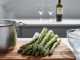 “Fresh green asparagus spears on a cutting board next to a pot of boiling salted water and a bowl of ice water, with wine glasses softly blurred in the background.”