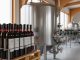 Stainless-steel fermentation tank and rows of commercial wine bottles in a modern winery under soft light.